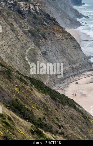 Sintra, Portugal - 24 June, 2020: Two people walking on remote beach below a staircase embedded into steep coastal cliffs on the Sintra Atlantic coast Stock Photo