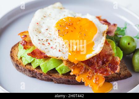 Keto breakfast concept. Fried egg with avocado and fried bacon on rye toast in gray plate. Stock Photo