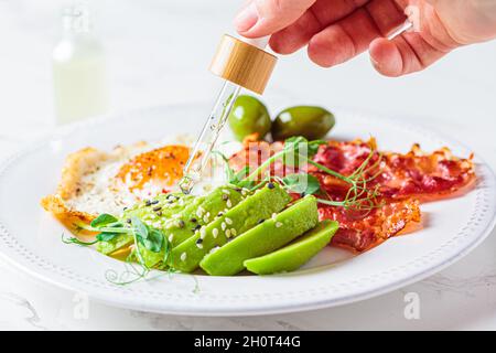 Cannabis essential oil CBD drip into food. Keto breakfast concept. Fried egg with avocado and fried bacon in a white plate. Stock Photo