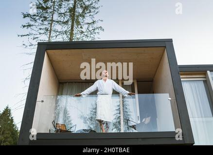 Resting Man dressed white bathrobe standing on forest house balcony and enjoying fresh air with nature forest pine trees view. Outside Scandinavian in Stock Photo