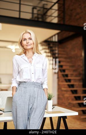 Stylish mature blonde business lady looking away while posing, standing in the office Stock Photo