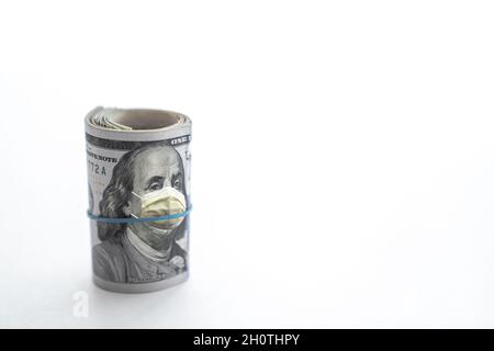 Rolled up wad of cash with hundred dollar bill on top and blue rubber band around the face of Benjamin Franklin wearing a yellow COVID-19 protective f Stock Photo