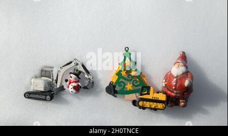 two models of toy excavators, souvenir snowman, figurine of Santa Claus, Christmas tree made of wood lie on white snow. concept for Christmas business Stock Photo