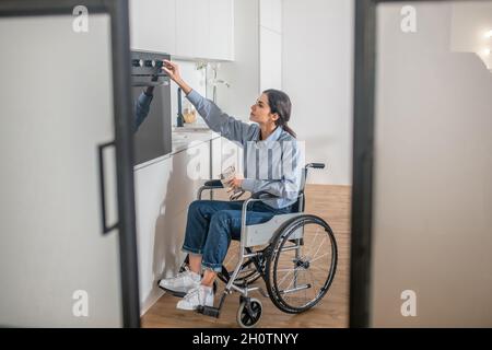 A girl on a wheelchair opening oven in the kitchen while cooking soemthing Stock Photo