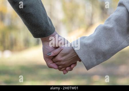Love, relationship, nature concept. Close-up shot of man and woman holds each other hands in blurred nature background during cold and sunny day Stock Photo