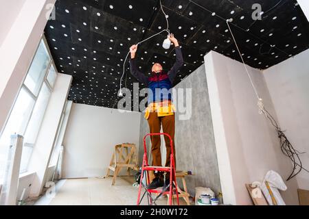 Full Length Portrait Of Electrician On Stepladder Installs Lighting To The Ceiling In Office Stock Photo