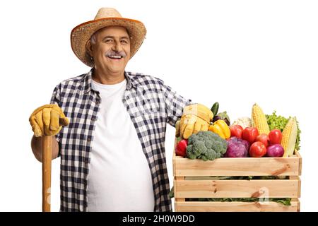 Mature farmer smiling and leaning on a crate of fresh organic vegetables with a shovel isolated on white background Stock Photo