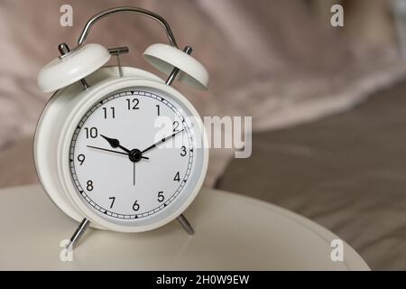 Small white retro alarm clock on a white table beside a bed with copy space Stock Photo