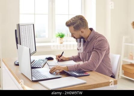Financial accountant writing something down while working on computers at his office desk Stock Photo