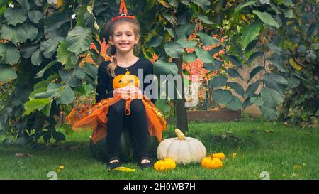 Little witch girl sitting on a pumpkin for Halloween. A girl in a witch costume hides under the umbrella elm tree and smiles. Children's trick or trea Stock Photo