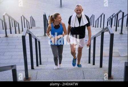 Sporty mature couple, man and woman in sportswear looking pleased, walking up the stairs after training together outdoors Stock Photo