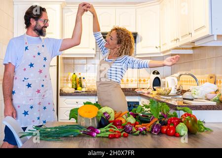 Happy adult couple dance and have fun together in the kitchen at home while preparing healthy vegetables on the table. Overjoyed woman and man in love Stock Photo
