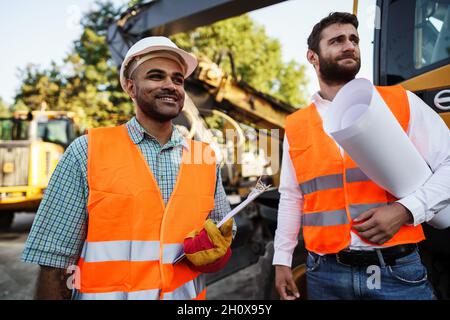 Two men engineers discussing their work standing against construction machines Stock Photo