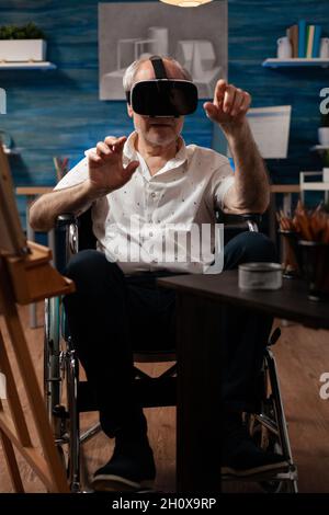 Invalid aged artist using vr glasses to draw masterpiece design on canvas and easel at fine art studio. Disabled senior man with modern headset sitting in wheelchair, for drawing hobby Stock Photo