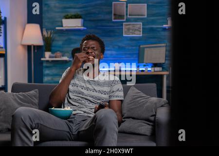 African american young adult laughing during comedy movie looking at television relaxing in living room. Black guy sitting on sofa while eating popcorn enjoying spending tine alone Stock Photo