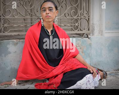 Handsome young nonbinary Latino wears a red Mexican rebozo and sits down on the sidewalk in front of a traditional wrought iron window grill. Stock Photo