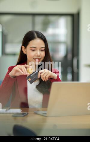 Young asian businesswoman sits in the office with a credit card in her hands, looking down at laptop screen. online shopping and payment concept Stock Photo