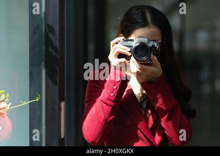 Young pretty female in a red suit with retro camera, taking picture, snap shot, focus the objects, hobby. Stock Photo