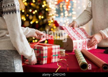 Man and woman preparing for Christmas. Couple in warm sweaters wrapping gifts. Decorated Christmas tree, boxes with presents, colourful wrapping paper Stock Photo