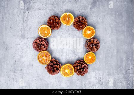 Modern Christmas wreath or Advent crown in bright colors: dried orange slices alternating with pine cones against a concrete-colored background. Stock Photo