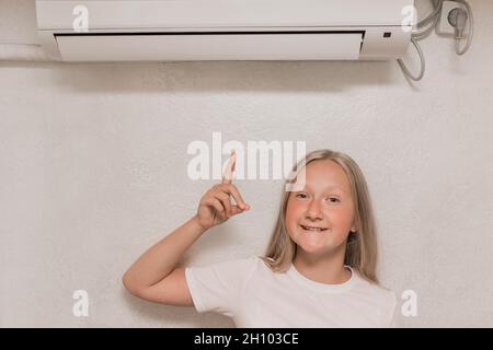Cute teenage girl blonde European appearance points a hand finger at the air conditioner on the wall in the room. Stock Photo