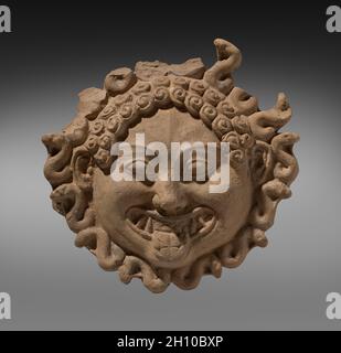 Gorgoneion Antefix (Ornamental Roof Tile), c. 500 BC. Greek/South Italian, Taranto. Terracotta (mold-made); height: 24.5 cm (9 5/8 in.).  This mold-made terracotta antefix, a type of ornamental roof tile, takes the form of a Gorgoneion, a disembodied Gorgon head. Nearly circular in shape, it features bulging, almond-shaped eyes and a grinning mouth with wide tongue and sharp teeth or tusks. S-shaped serpents surround the head, some standing tall. Traces of pigment suggest a more vivid original appearance, perhaps considered both terrifying and protective. Stock Photo