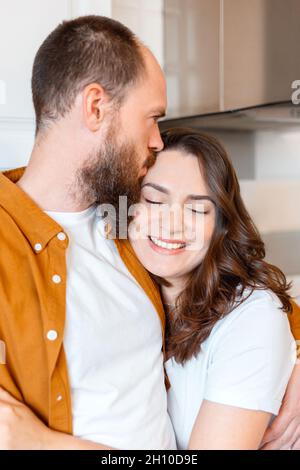 Happy family couple portrait Husband kisses his wife on forehead in kitchen. Couple of young middle aged man and Loving wife woman smiling with closed Stock Photo