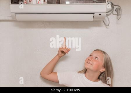 Cute teenage girl blonde European appearance points a hand finger at the air conditioner on the wall in the room. Stock Photo
