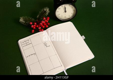 High angle view of an opened page of notepad with plans for day, alarm clock and snowy branch of holly Christmas plant isolated on green background Stock Photo