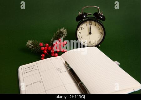Close-up of an opened page of notebook, agenda with plans for day, alarm clock and snowy branch of holly Christmas plant isolated on green background Stock Photo