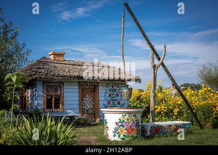 Zalipie, Poland - August 1, 2021: An old white wooden cottage and a traditional well, painted in floral pattern. Yellow flowers in the garden. Stock Photo