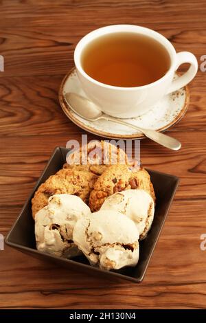 Oatmeal cookies with walnuts and raisins, meringue cookies with hazelnuts in a square bowl and a cup of green tea on a wooden table. Closeup Stock Photo
