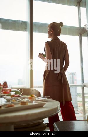mature asian woman looking at view through window of hotel room with breakfast on table Stock Photo