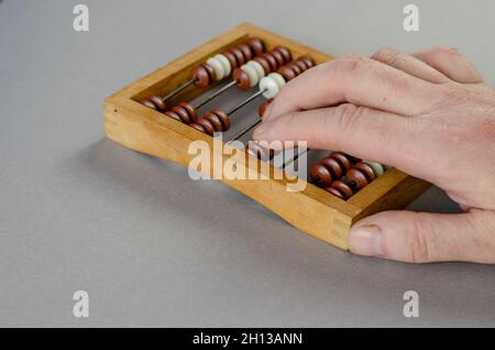 An accountant makes calculations on wooden abacus. The right hand of a middle-aged man moves wooden beads. Mechanical mathematical device on gray back Stock Photo