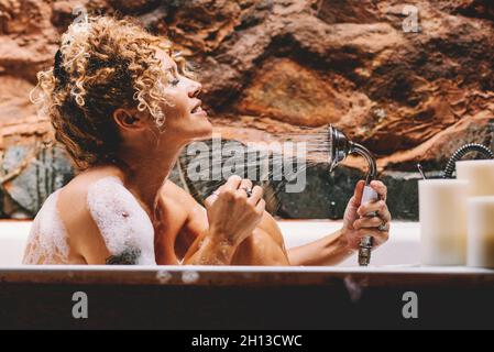 Happy beautiful young adult woman washing with shower sitting in the bathroom at home. Body care and beauty make over lifestyle female people enjoy so Stock Photo