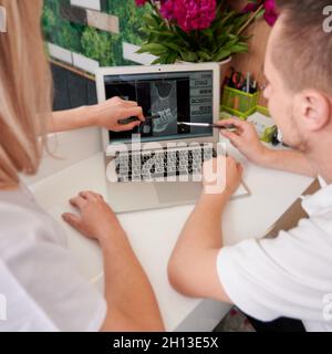 Back view on young female dentist with assistant analyzing x ray image on pc in clinic. Working process. Focus on the laptop. Woman pointing on computer screen. Dentistry concept Stock Photo