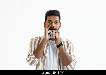 Shocked young Indian man holding hands behind head and keeping mouth open while standing against white background Stock Photo