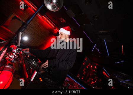 New Year in night club. Merry Christmas Young confident DJ in red santa claus hat mixing music on turntables at holiday party. Shining disco ball Stock Photo