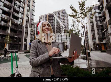 Blonde woman in santa's hat wearing casual clothes sitting outside and working on her laptop. Merry Christmas and Happy New Year Young freelancer Stock Photo
