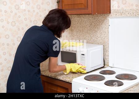 Brunette housewife yellow rubber gloves washes the kitchen microwave oven. Stock Photo