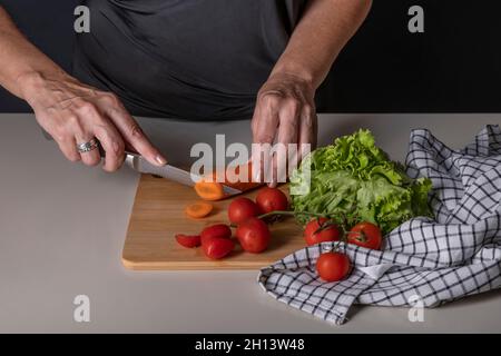 Female hands cut a carrot on a wooden cutting board, to prepare a salad with red cherry tomatoes Stock Photo