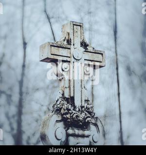 A beautiful white old stone cross covered with black mold stands in the cemetery in cloudy gray weather. Grief and sorrow. Stock Photo