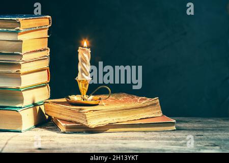 Vintage background with a stack of old antique books and lighten candle in a golden candlestick at night. Spooky dark academia atmosphere. Cozy fall s Stock Photo