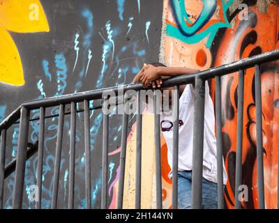 Medellin, Antioquia  Colombia - January 6 2021: Afro American Boy Lays on a Black Metal Fence Sadly against Mural Background in Comuna 13 Neighborhood Stock Photo