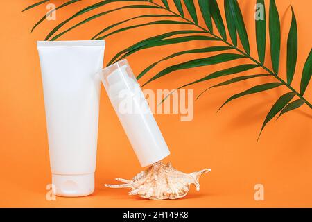 Two white blank cosmetics bottles with sunscreen, suncream or other cosmetic product, seashell and green brunch palm on orange background. Concept ski Stock Photo