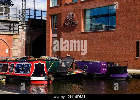 Sunlit narrow boats moored side by side on water, city centre Hilton hotel & old railway tunnel - Leeds Liverpool Canal, West Yorkshire, England, UK. Stock Photo