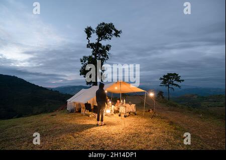 Young man standing and holding lantern shine with friends sitting in large tent on hill in campsite at countryside. Stock Photo