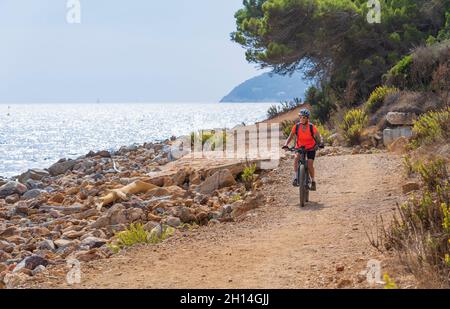 nice woman riding her electric mountain bike at the coastline of mediterranean sea on the Island of Elba in the tuscan Archipelago, in front of Porto Stock Photo