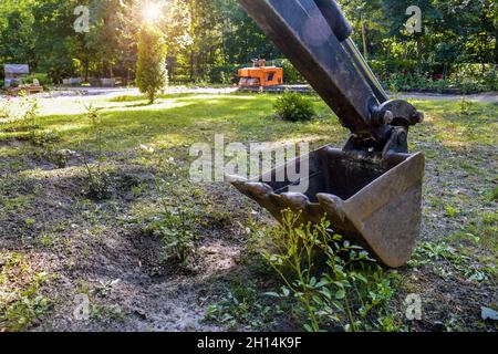 Bucket of small excavator is located on lawn. Strong teeth are ready to bite into ground. Construction work in park. Close-up. Selective focus. Stock Photo