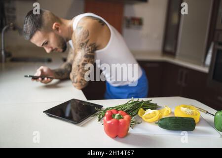 Young fit male using his smartphone while cooking healthy food in the kitchen Stock Photo